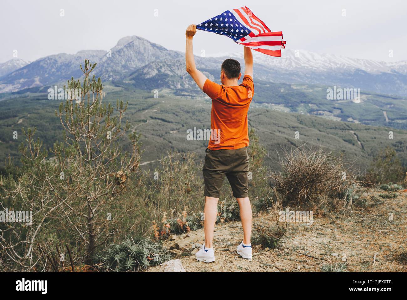 Young man standing on a rock cliff and waving the US flag while looking at mountains in the background. Male traveller waving American flag standing Stock Photo