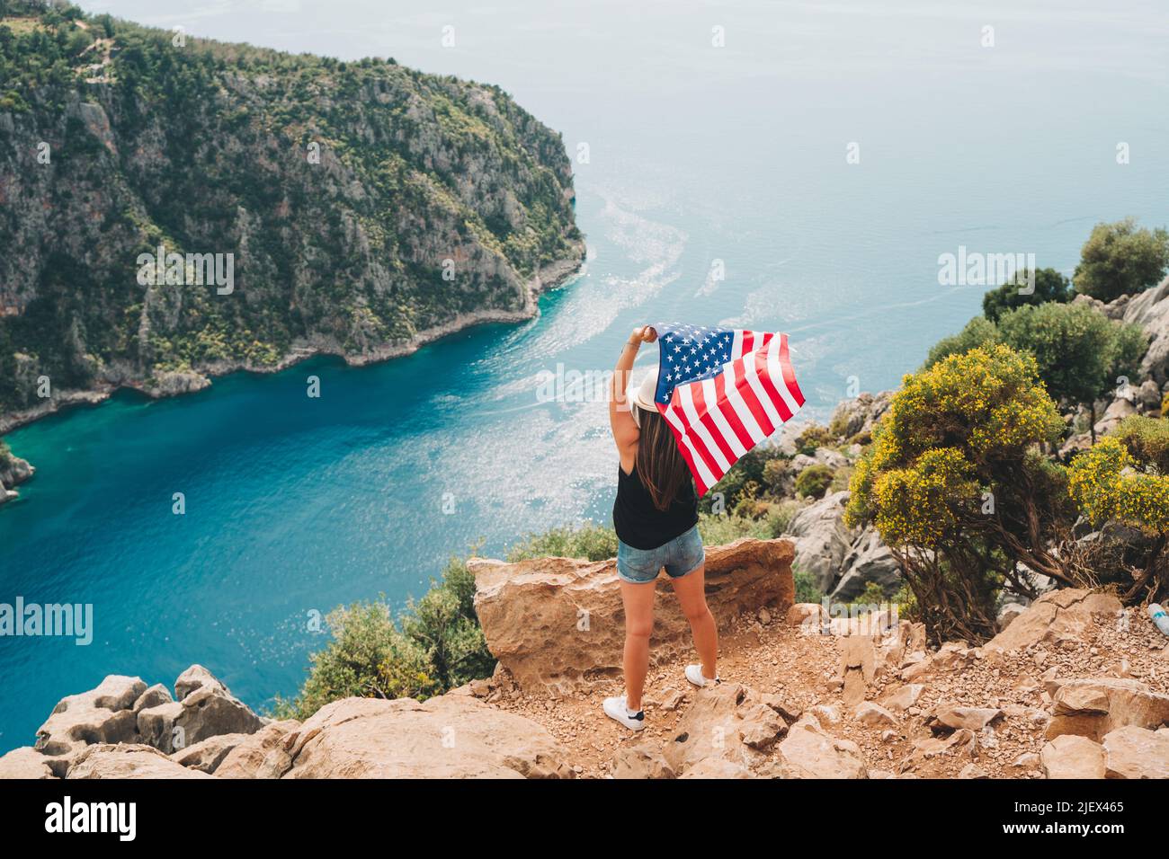 Young woman standing on a rock cliff and waving the US flag while looking at sea beneath. Girl traveller waving the American flag while standing on a Stock Photo