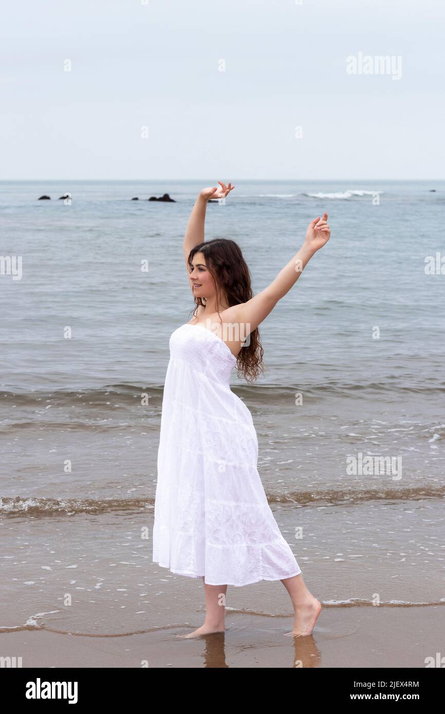 young brunette woman raising her arms on the beach in a white dress ...