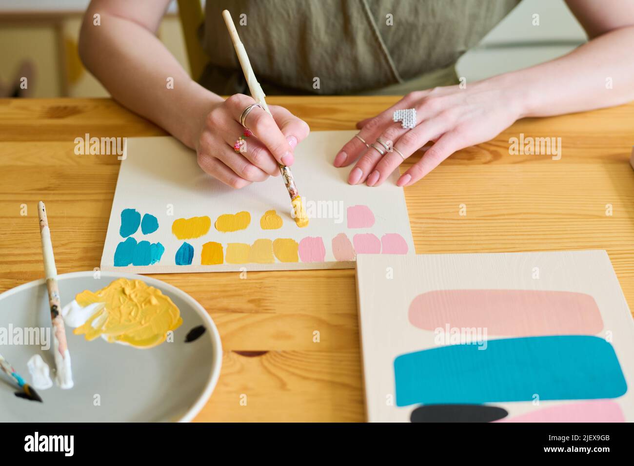 Hands of young woman with paintbrush making color swatches for her new painting while sitting by wooden table in workshop Stock Photo