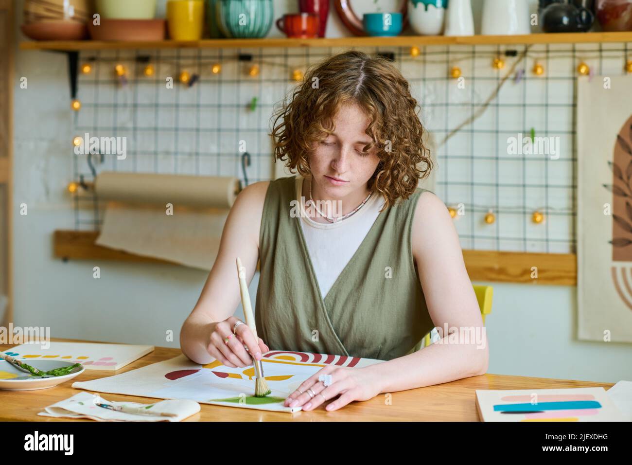 Young modern female in casualwear sitting by table and working over creation of new abstract painting in workshop or studio Stock Photo