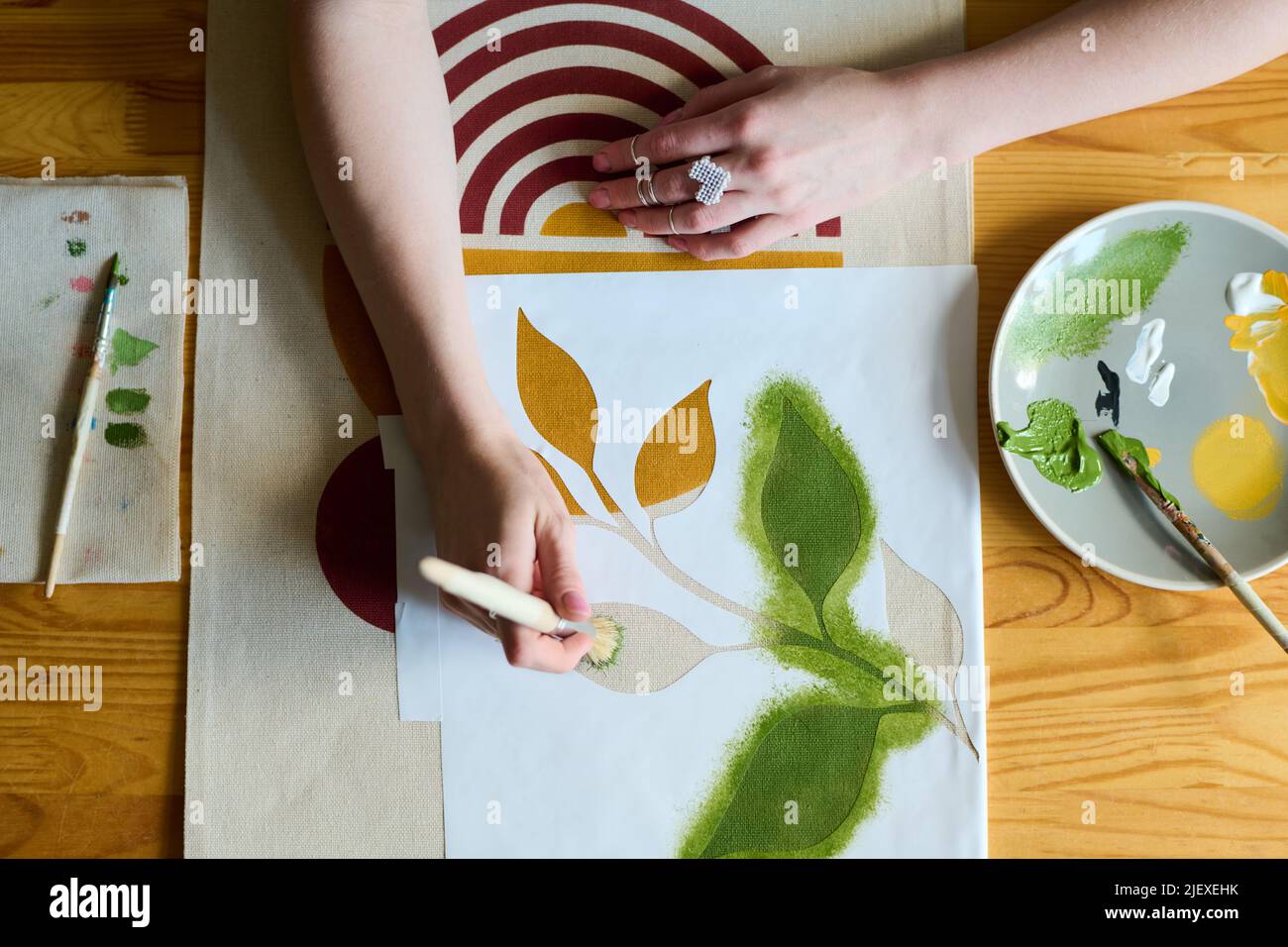 Above angle of hands of young female artist with paintbrush painting green leaves on paper while sitting by table in workshop Stock Photo