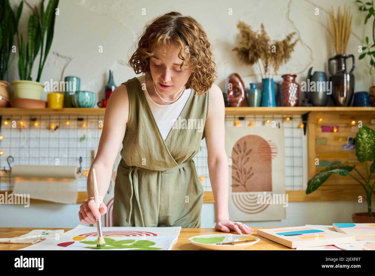 Young creative female bending over table while painting abstract picture on paper with paintbrush and green watercolor in workshop Stock Photo