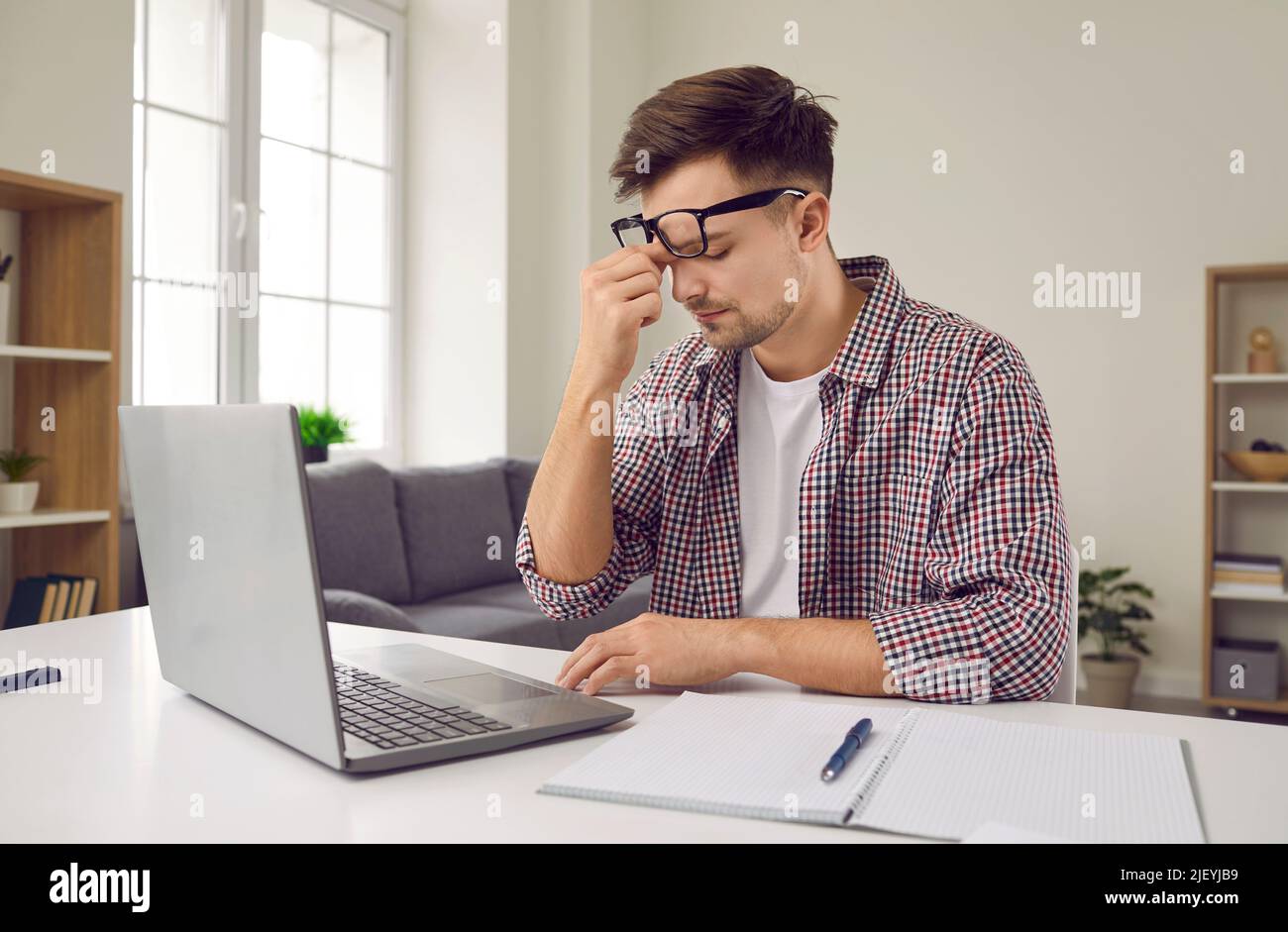 Sad tired overworked young man working on laptop rubs between eyes trying to remember something. Stock Photo