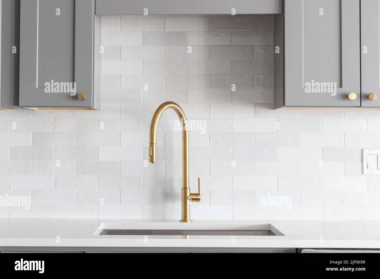 A kitchen sink detail shot with a gold faucet, marble backsplash, grey cabinets, and gold hardware. Stock Photo
