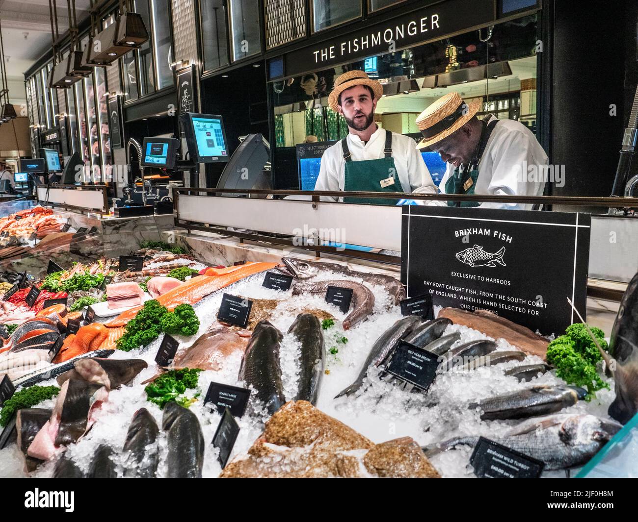 Harrods Fishmonger Fresh Brixham Fish Food Hall interior featuring 'The Fishmonger' display with a wide variety of exclusive day boat hand picked Brixham UK British fish. Seafood crustacea on display for sale. Sales assistants wearing traditional straw boater hats, preparing a customers selection, Harrods Food Hall Fishmonger department Store Mall Shop,  Brompton Rd, Knightsbridge, London SW1X 7XL Stock Photo