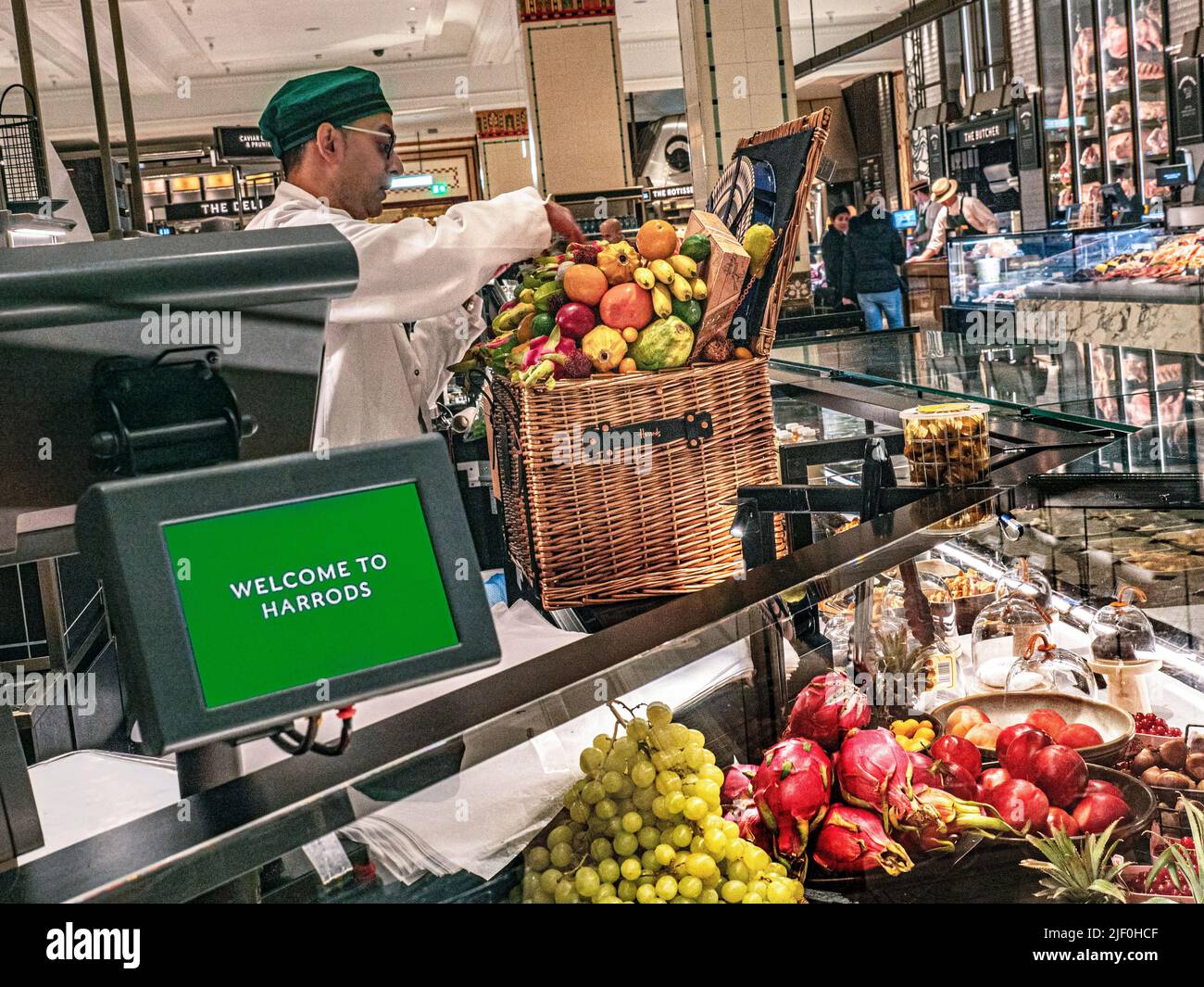 Harrods Food Hall interior with sales assistant preparing a fresh fruit Harrods hamper basket display 'Welcome to Harrods' weighing machine and wrapping point in foreground. Harrods Food Hall, Mall,  Brompton Rd, Knightsbridge, London SW1X 7XL Stock Photo