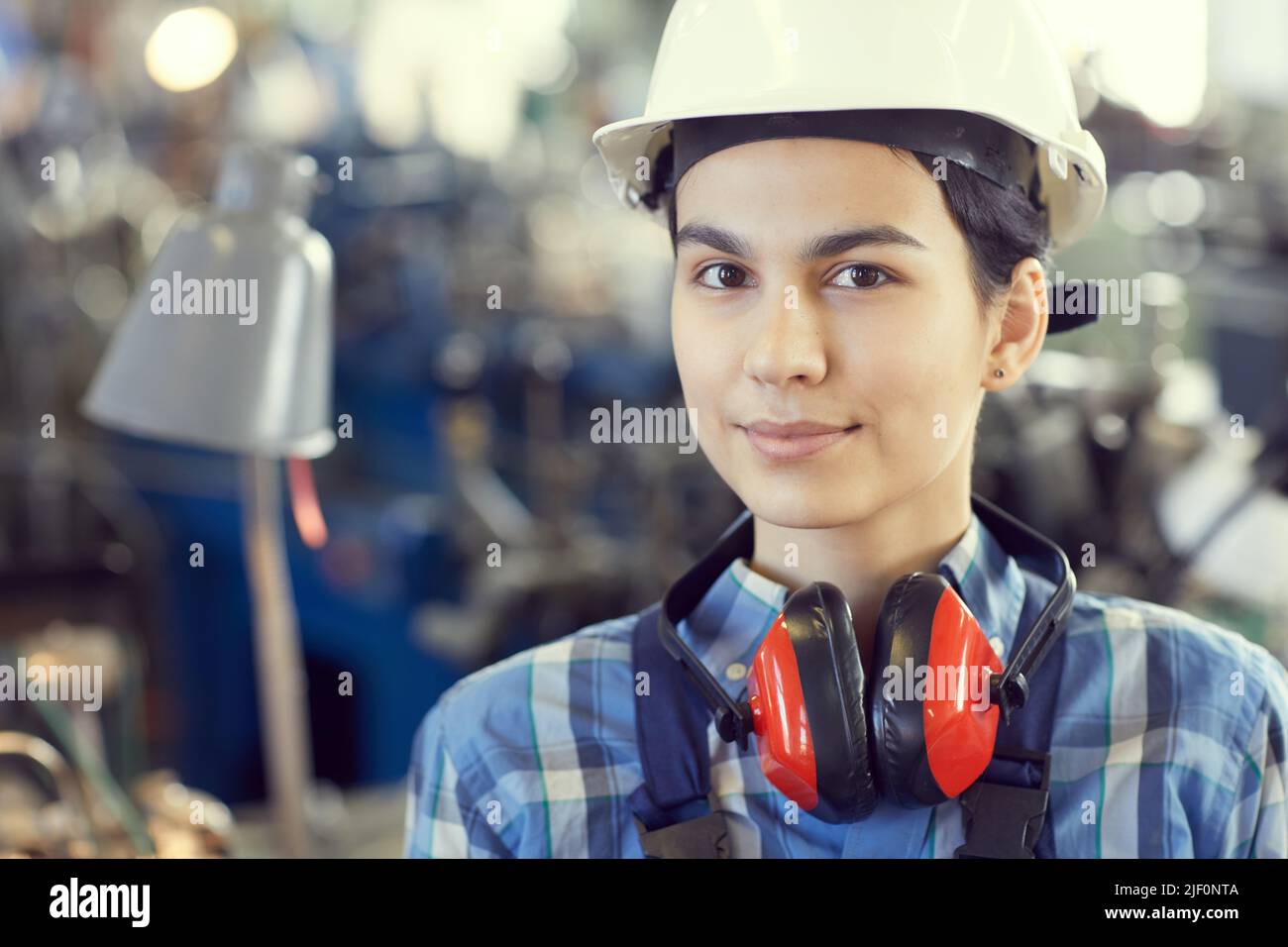 Portrait of smiling tomboy wearing hardhat and ear protectors standing at factory shop Stock Photo