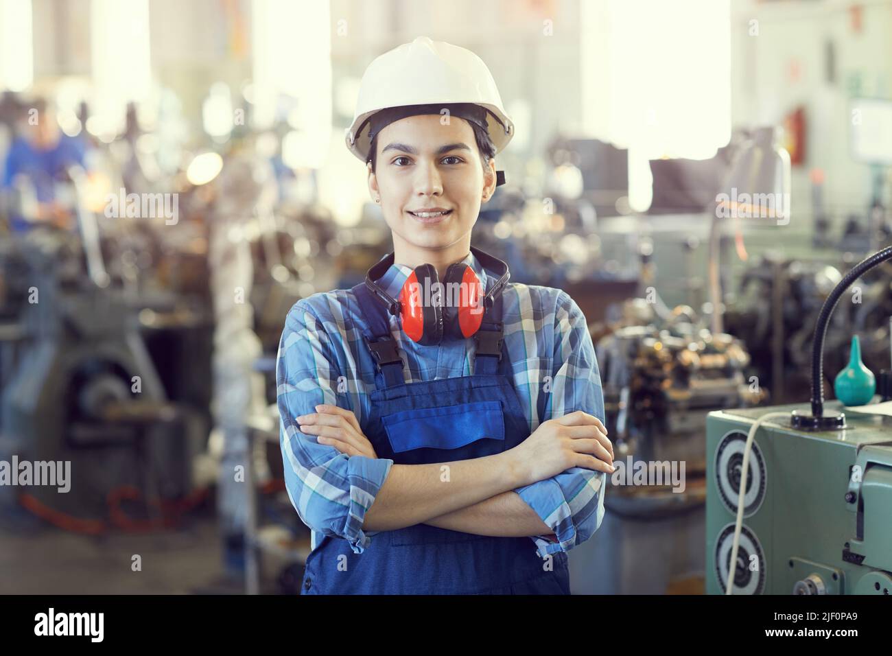 Portrait of positive confident young female factory engineer in hardhat and ear protectors around neck crossing arms on chest and standing in industri Stock Photo