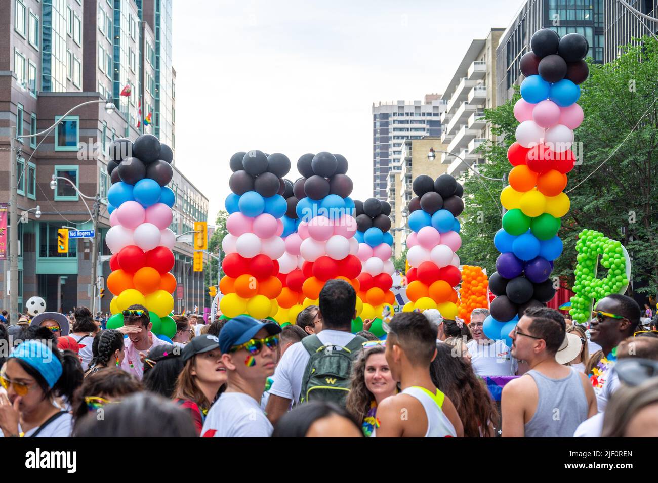 Crowd of people marching in Bloor Street during Pride Parade. They are holding columns of balloons with rainbow colors. Stock Photo