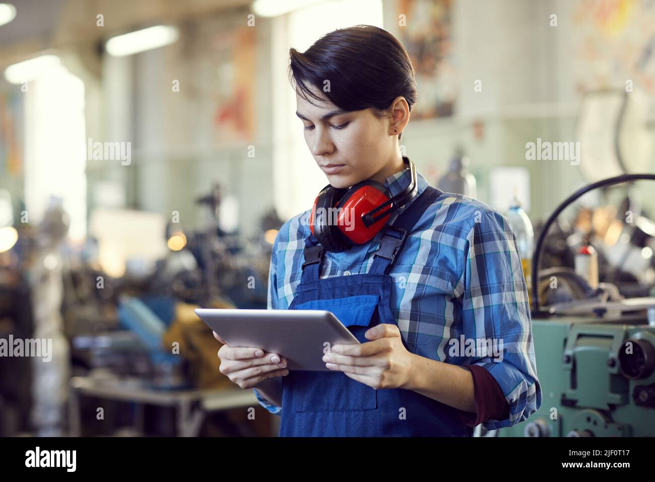 Serious busy female factory engineer with short black hair standing in workshop and using tablet for lathe examination Stock Photo