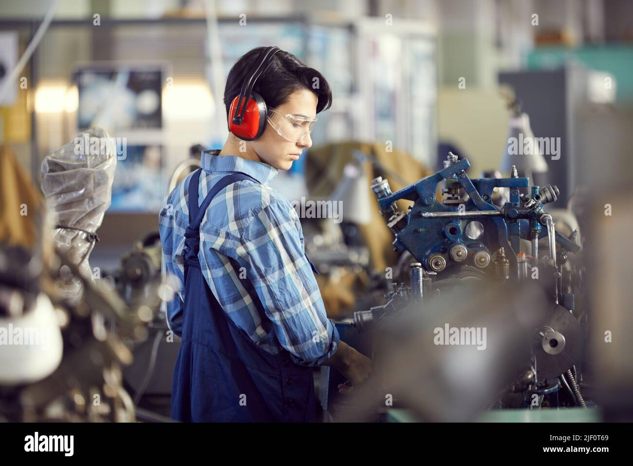 Serious rough girl in overall and ear protectors standing in industrial shop and examining factory machine before work Stock Photo