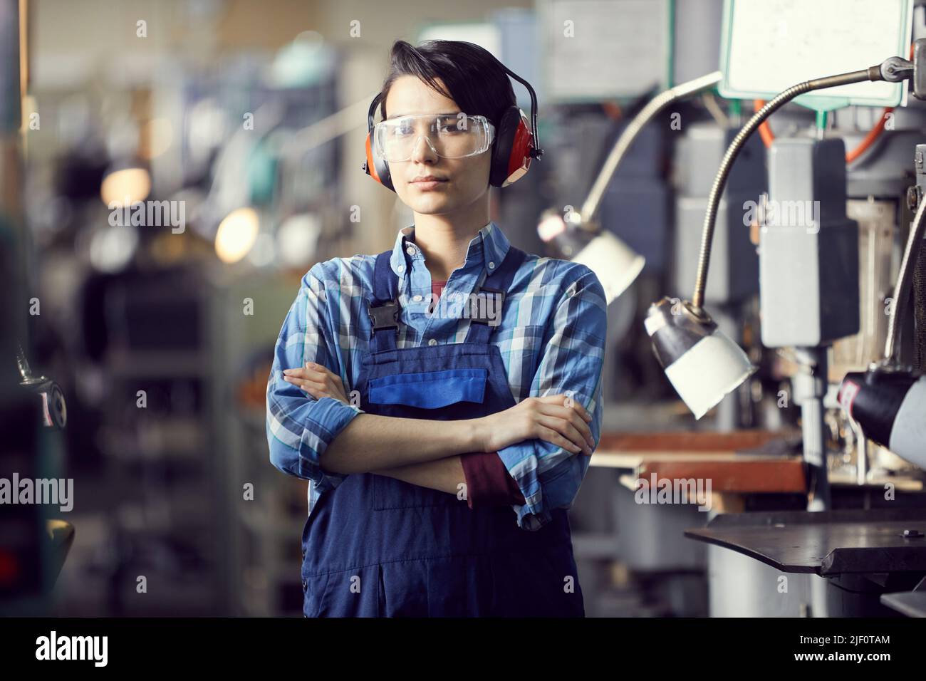 Portrait of serious professional female engineer in ear protectors and safety goggles standing at factory and crossing arms on chest Stock Photo
