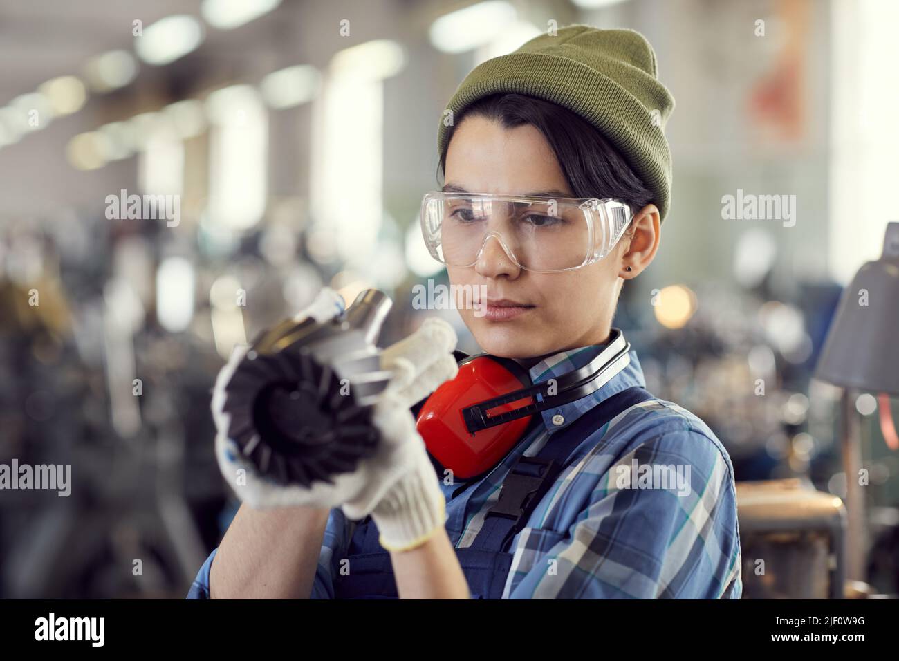 Busy female repair engineer in beanie hat and safety goggles looking at machine tool while finding out breakdown of lathe Stock Photo