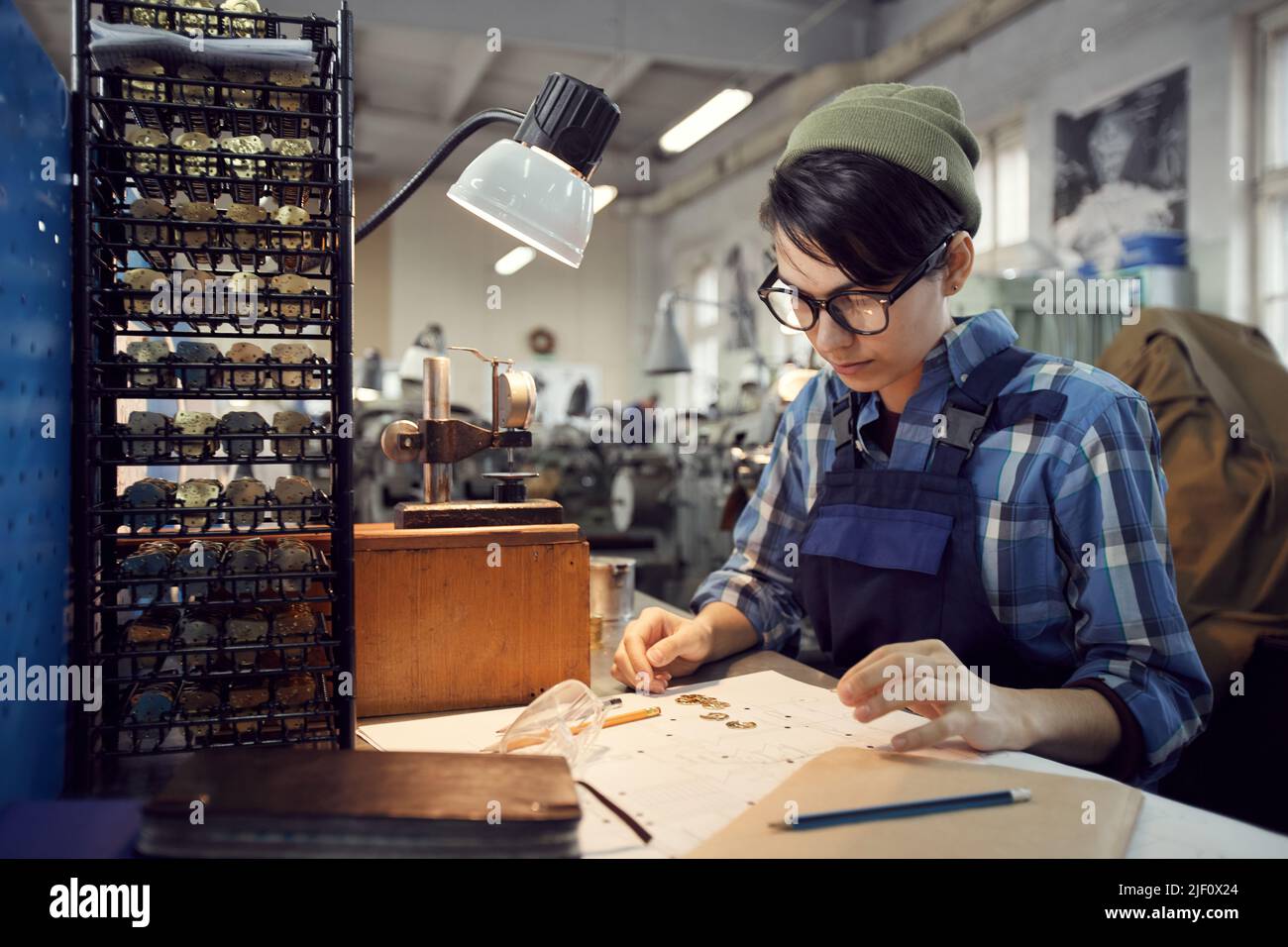 Serious hipster female engineer in beanie hat sitting at desk with sketch and watch plates and examining produced watch movements Stock Photo