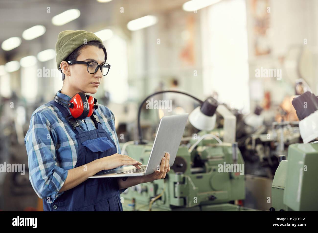 Serious focused young female industrial engineer in beanie hat and eyewear standing in factory shop full of lathes and using laptop Stock Photo