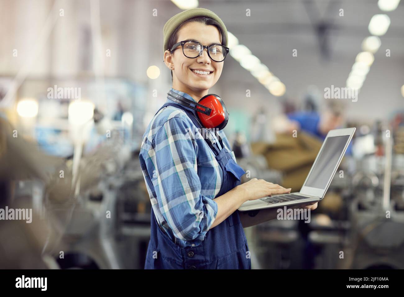 Portrait of cheerful confident hipster female maintenance engineer with ear protectors around neck using laptop at industrial factory Stock Photo