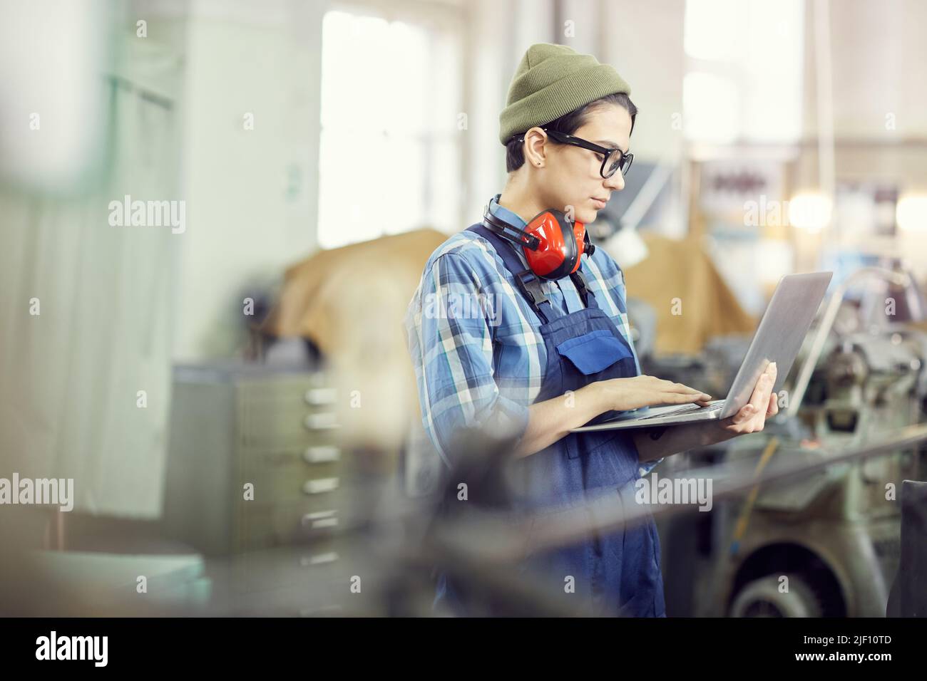 Serious busy young female engineer in hipster hat standing in factory shop and identifying industrial machine errors using laptop Stock Photo