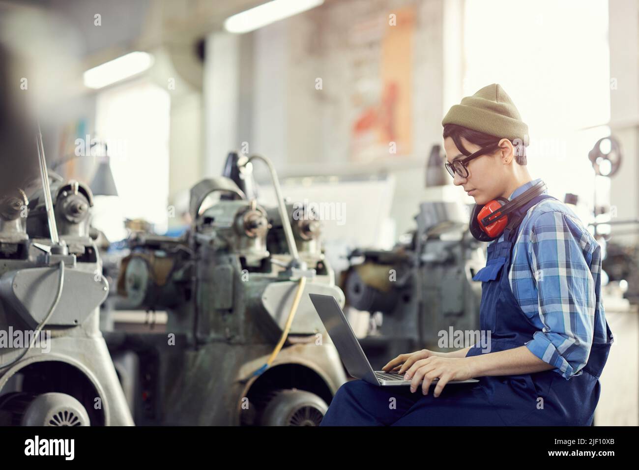 Concentrated young female production engineer in khaki beanie hat and blue overall sitting in workshop with lathes in background and analyzing sketch Stock Photo