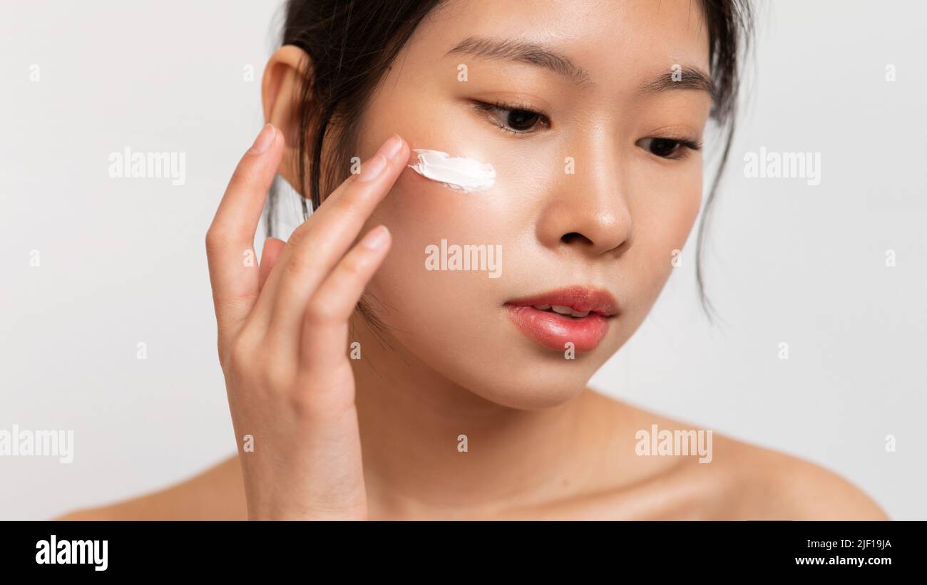 Closeup of asian lady applying anti-aging or moisturising face cream on cheek, standing on white studio background Stock Photo