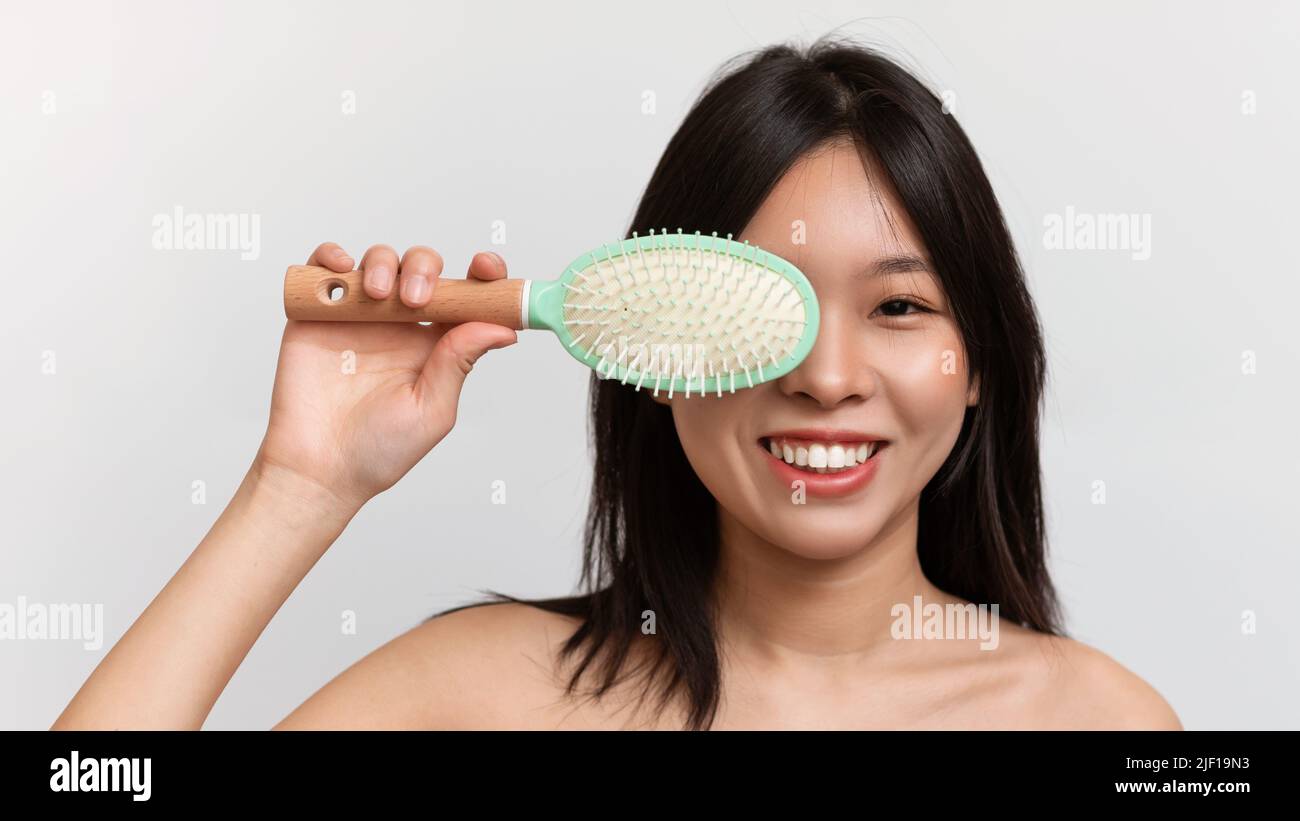 Happy japanese lady holding hair brush, covering eye and smiling at camera, posing over white background, panorama Stock Photo