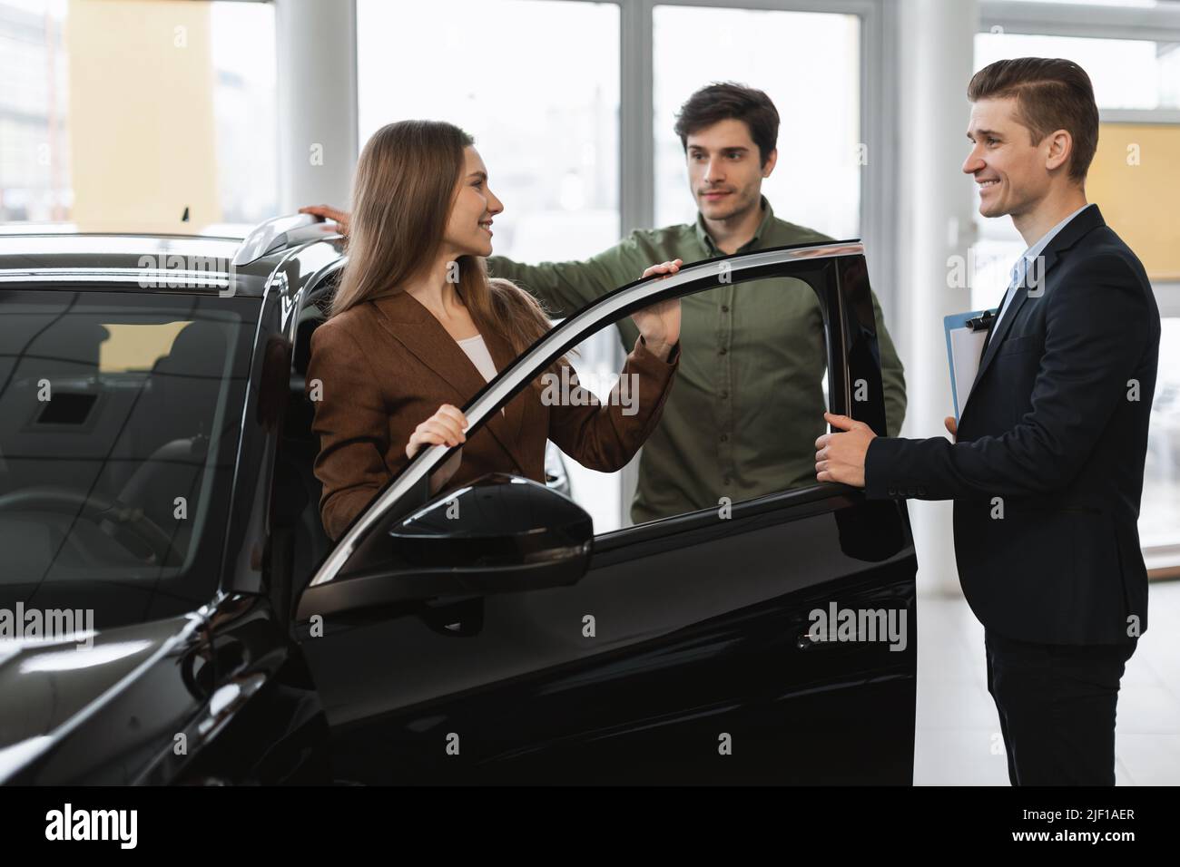 Positive young couple discussing purchase or lease of new car with auto salesman at modern dealership Stock Photo