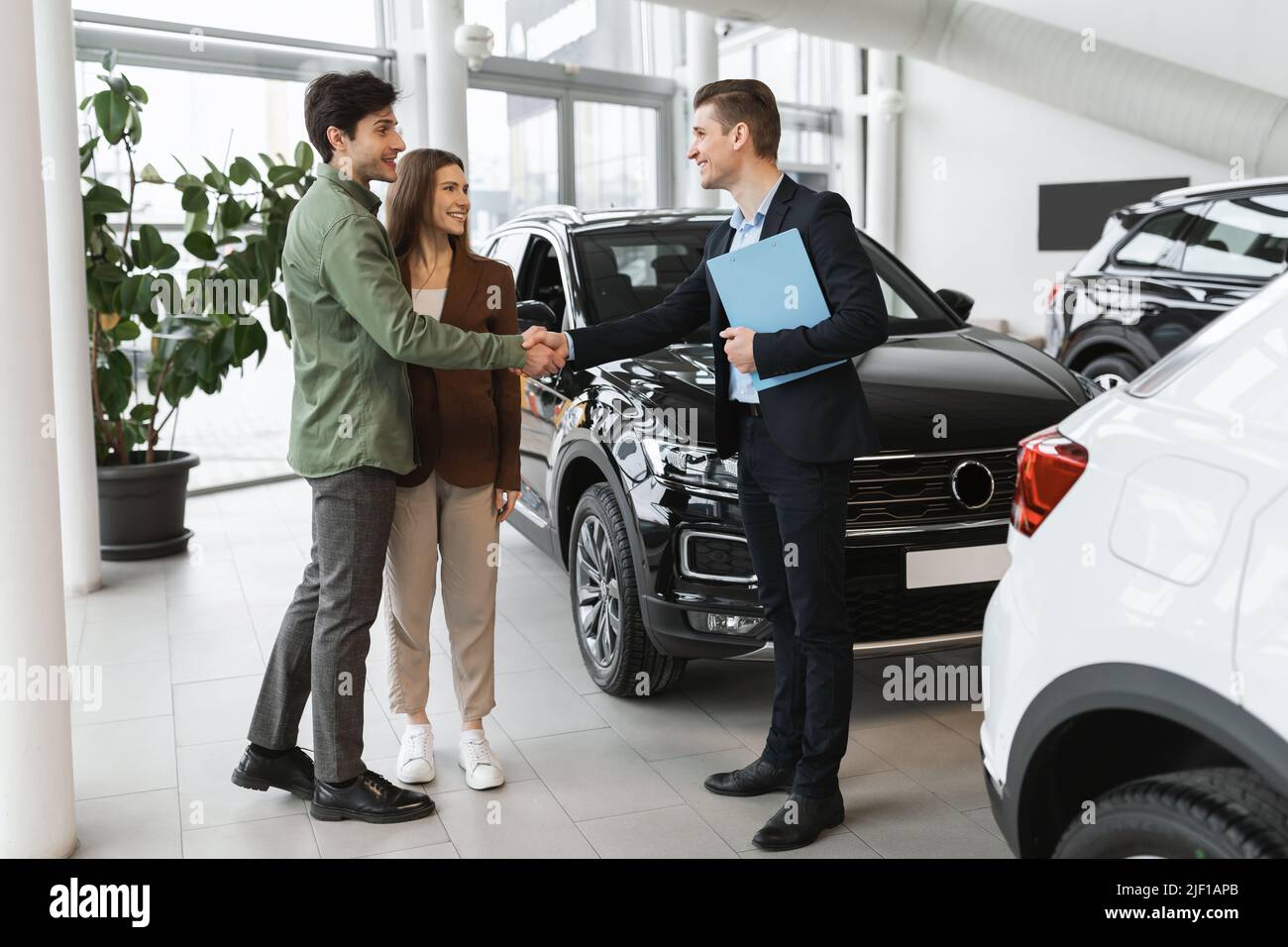 Young family making deal, buying or renting modern automobile, shaking hands with car salesman at auto showroom Stock Photo