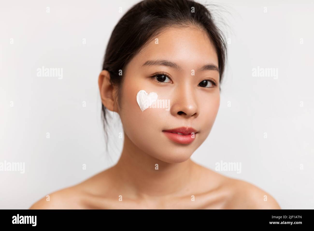 Closeup portrait of pretty korean lady with moisturizer cream on her face in heart shape, white studio background Stock Photo