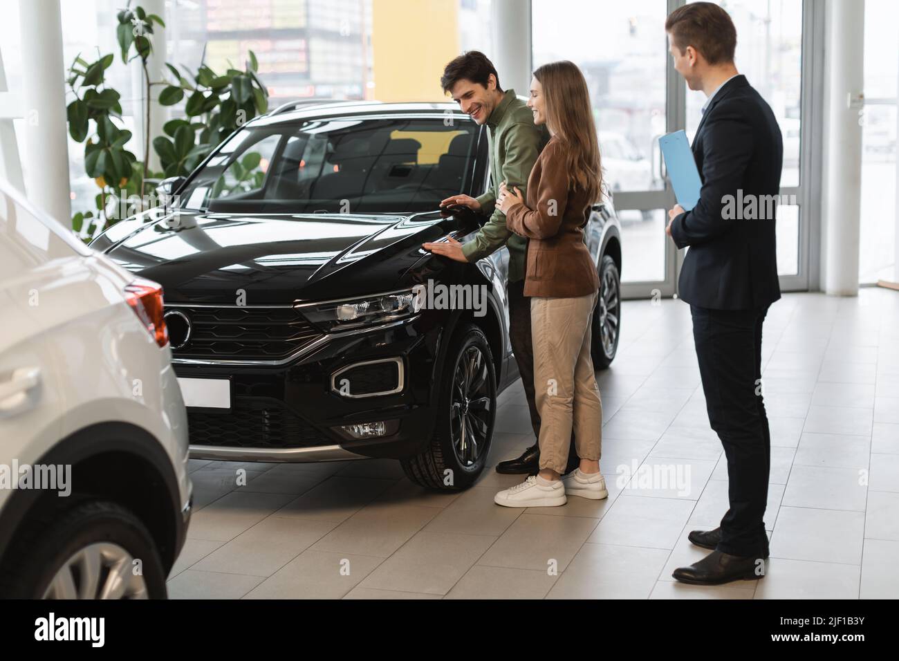 Cheery millennial couple speaking to car salesman about buying or renting new vehicle at dealership, copy space Stock Photo