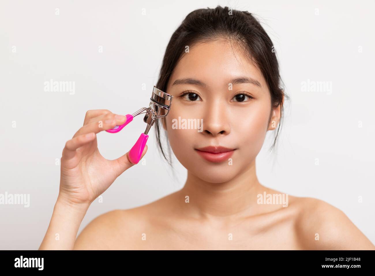 Closeup portrait of pretty asian lady using eye lash curler on long eyelashes, posing on white