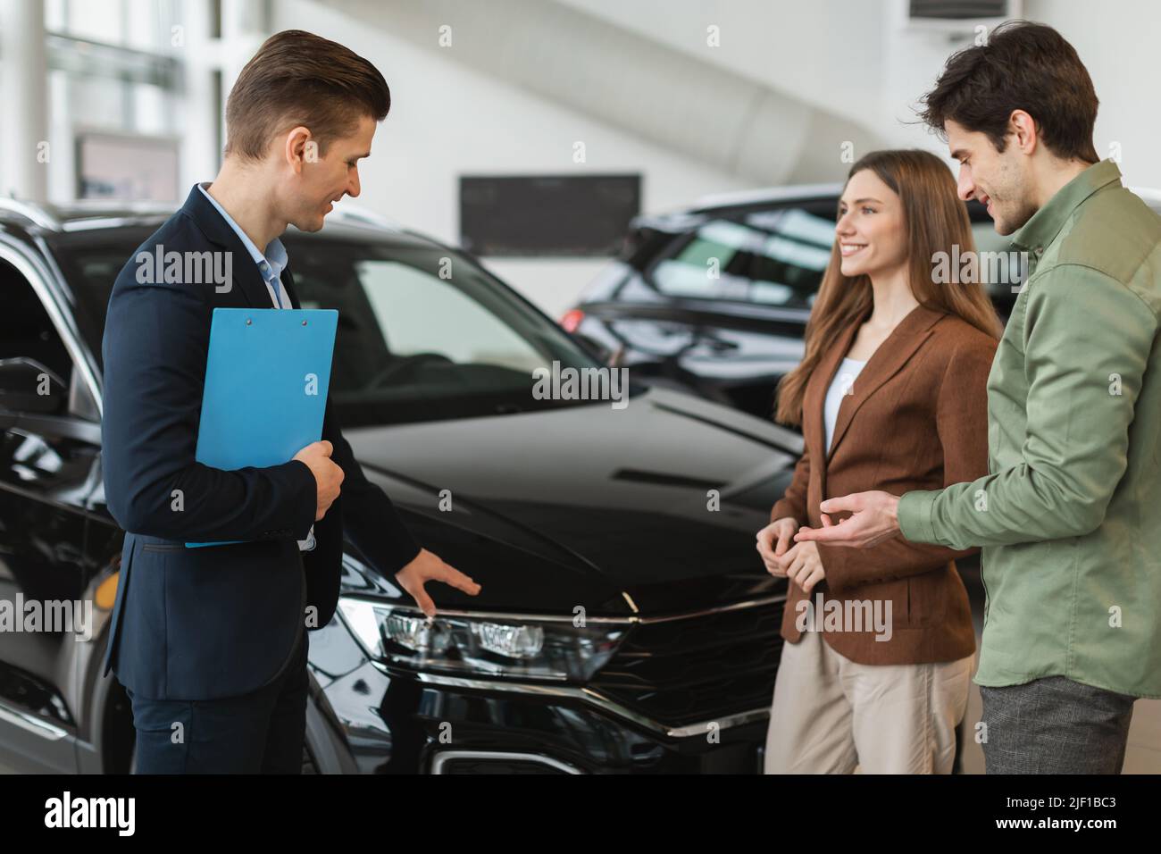 Positive young spouses speaking to salesman about purchasing new auto at dealership shop. Car purchase or rental Stock Photo
