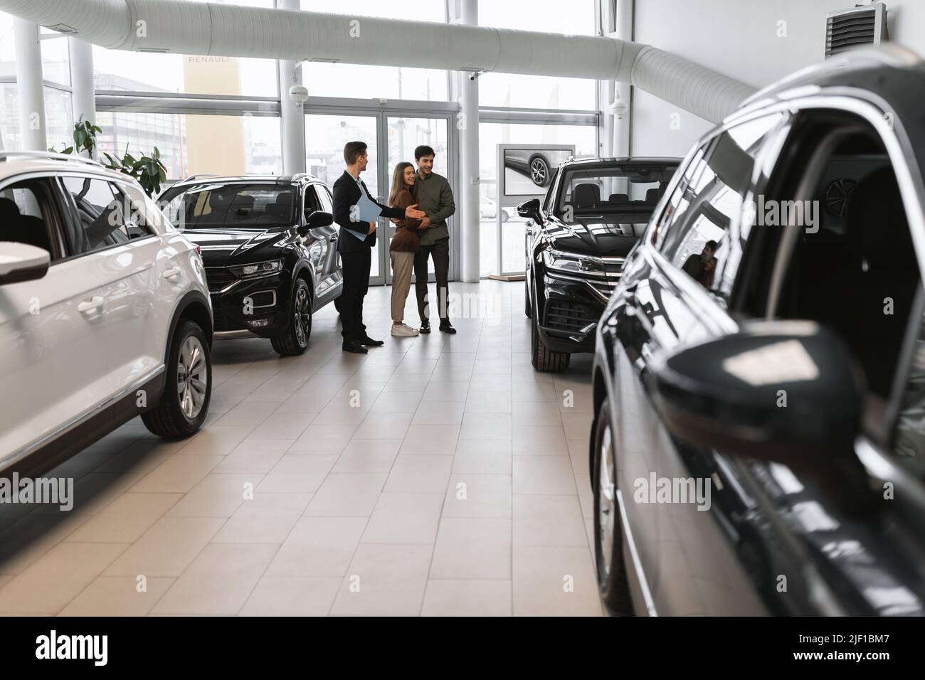 Full length of millennial couple choosing new car, having discussion with auto salesman at vehicle dealership Stock Photo