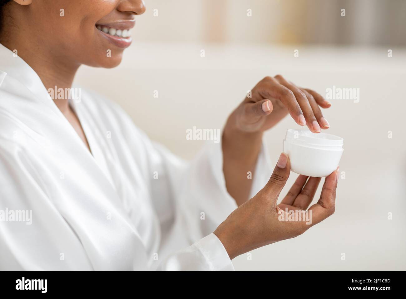 Smiling black woman holding open jar with moisturising cream, making beauty treatments Stock Photo