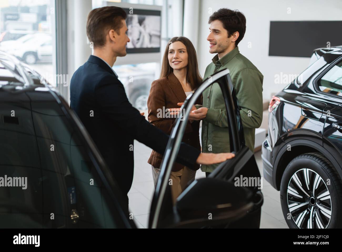 Joyful young couple consulting with auto salesman, picking new car at automobile dealership Stock Photo