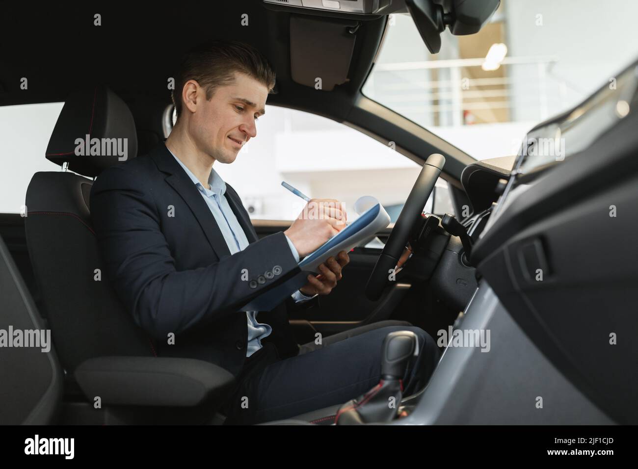 Positive young auto salesman sitting inside new car, making checkup, taking notes during test drive at dealership Stock Photo