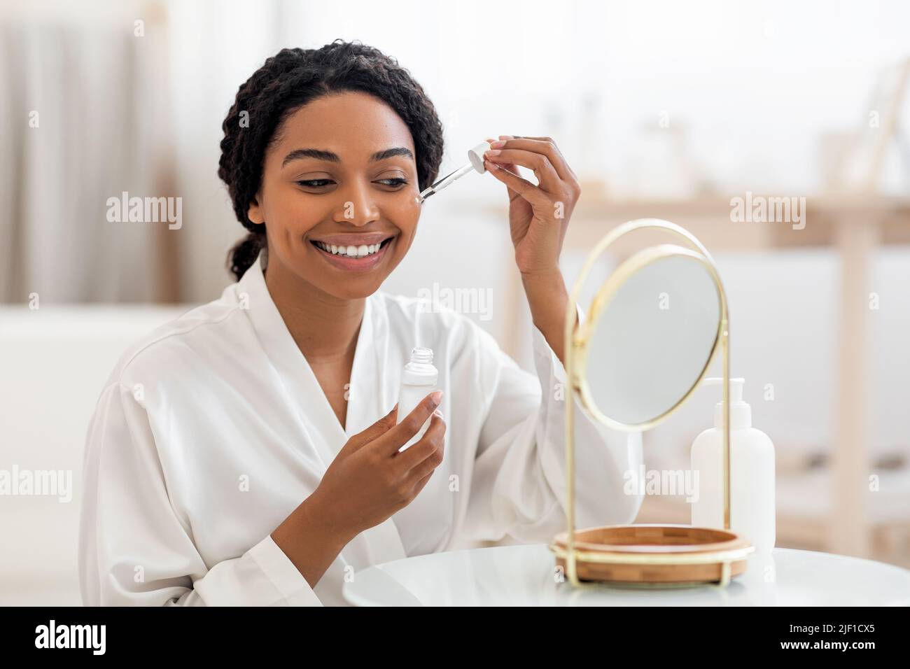 Skincare Concept. Beautiful Black Lady Applying Face Serum Near Mirror At Home Stock Photo