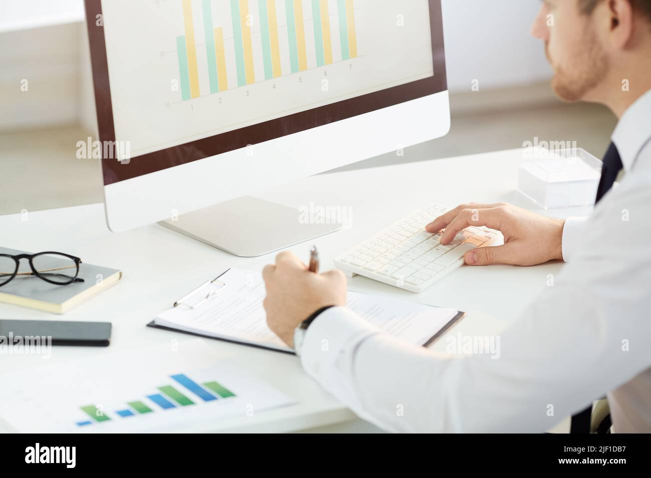 Over shoulder view of busy young market analyst using modern computer and making notes in report while analyzing graph Stock Photo