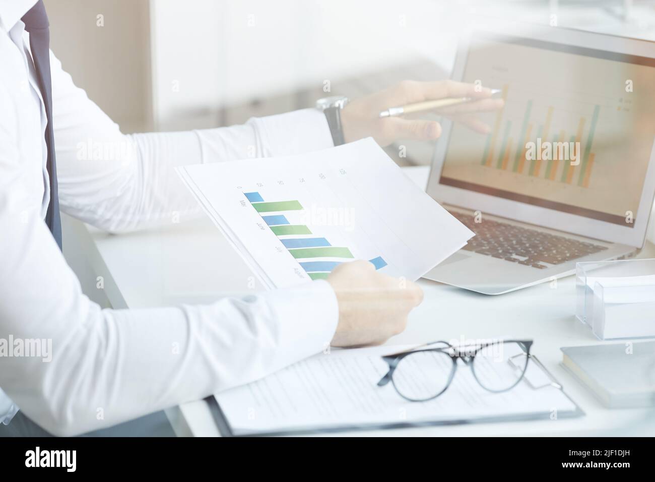 Close-up of unrecognizable market analyst sitting at desk and comparing graphs in laptop and papers while working in office, behind glass view Stock Photo