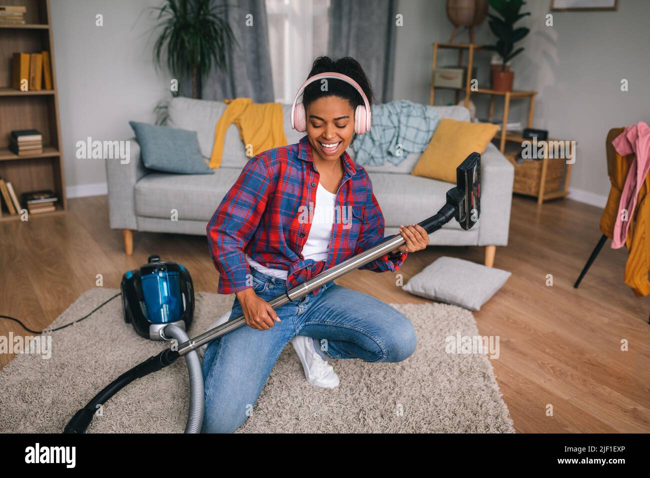 Cheerful young african american female in headphones playing at imaginary guitar as vacuum cleaner in living room Stock Photo