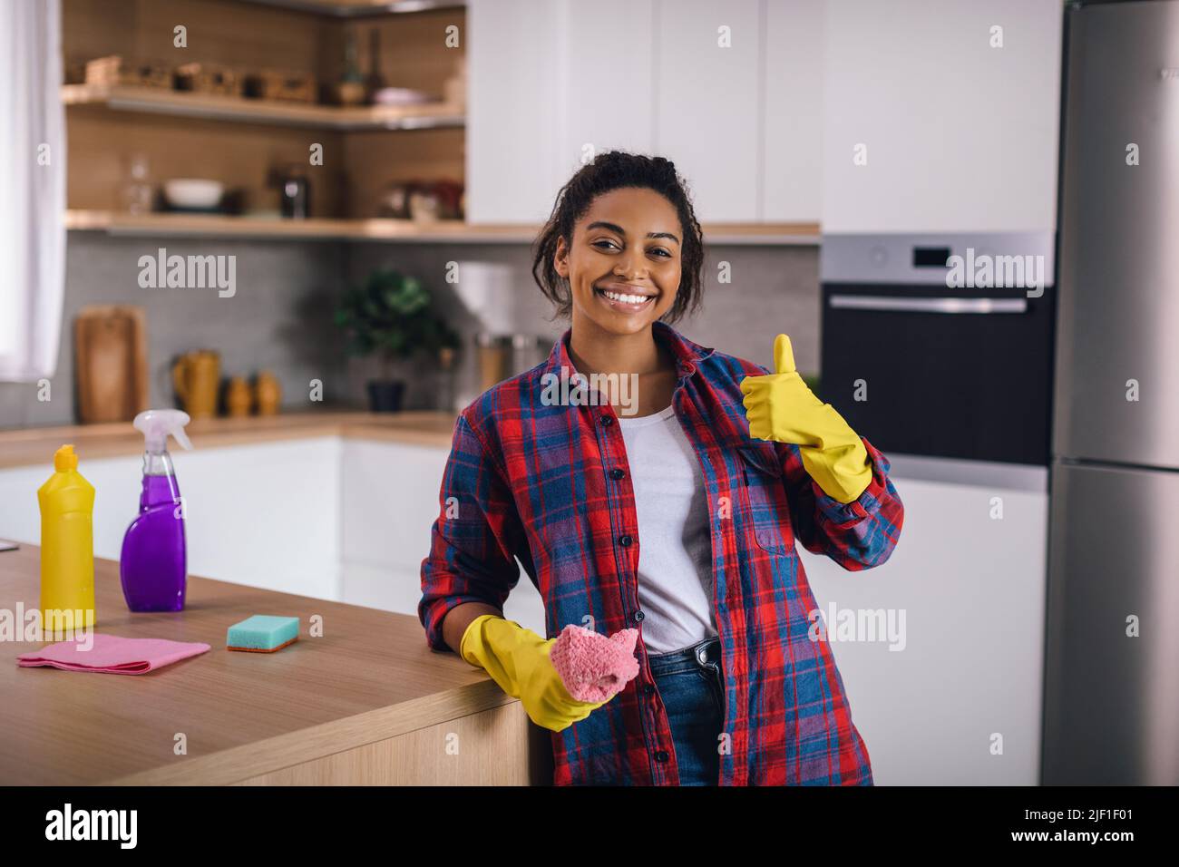 Cheerful millennial african american female in rubber gloves with rag and cleaning supplies showing thumb up Stock Photo