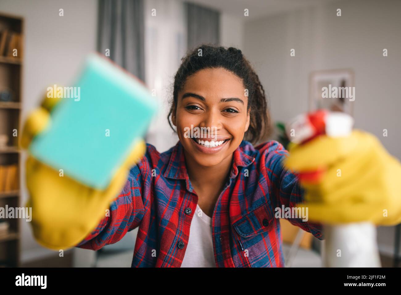 Smiling young african american lady in rubber gloves with spray and sponge wipes camera at modern kitchen Stock Photo