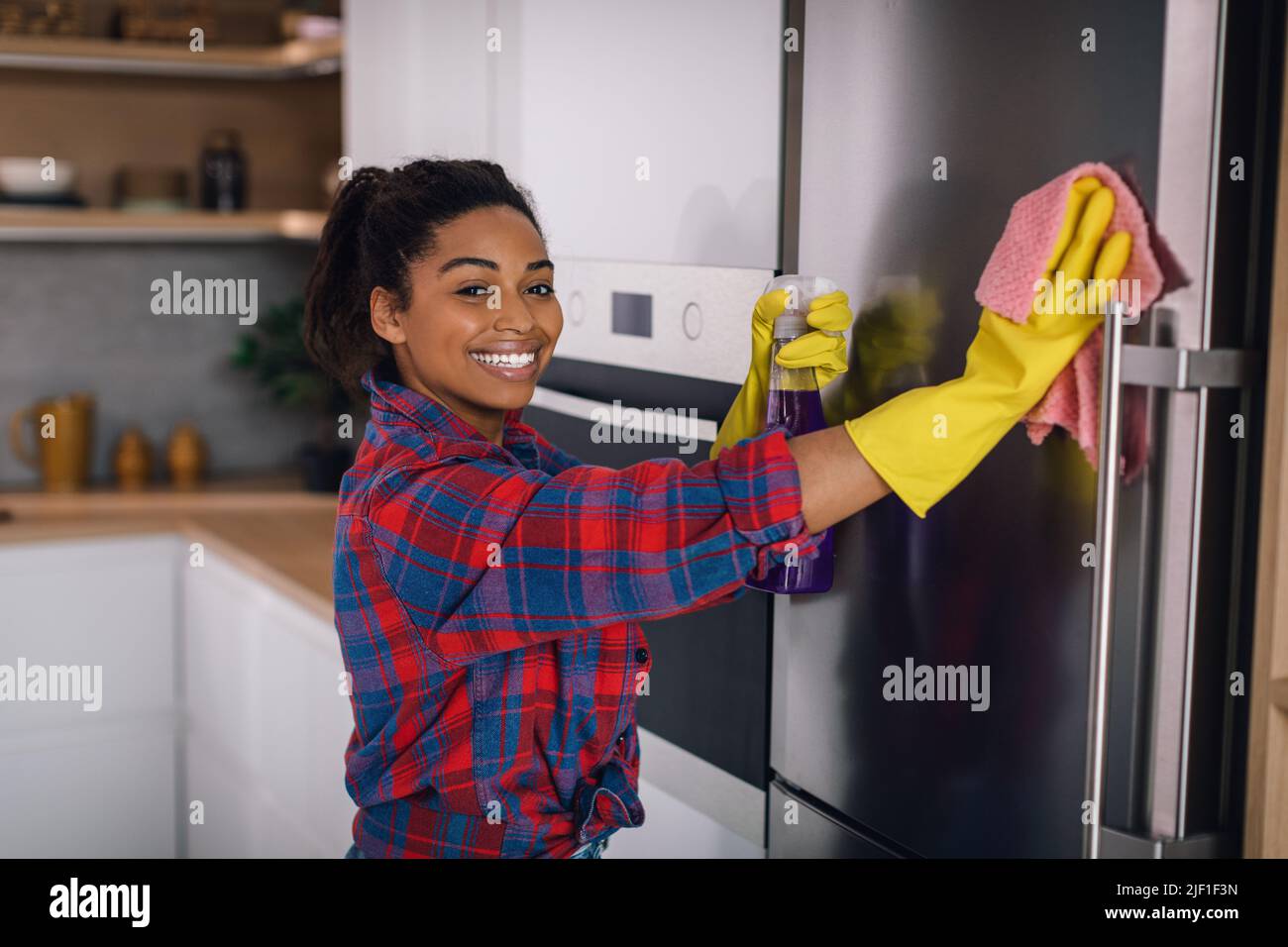 Happy young african american woman in rubber gloves wipes refrigerator with spray and sponge Stock Photo