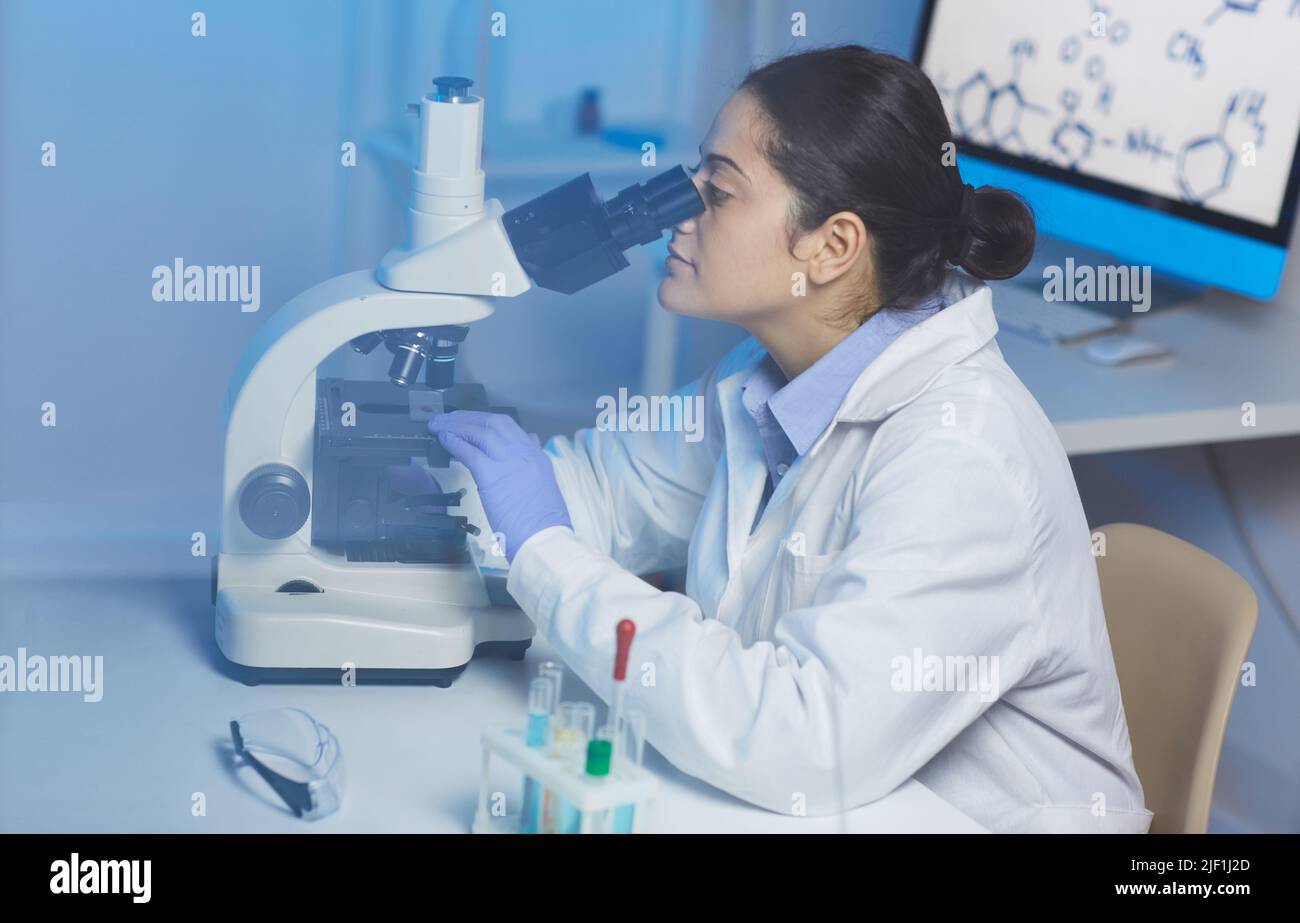 Concentrated young Indian female lab technician in white coat sitting