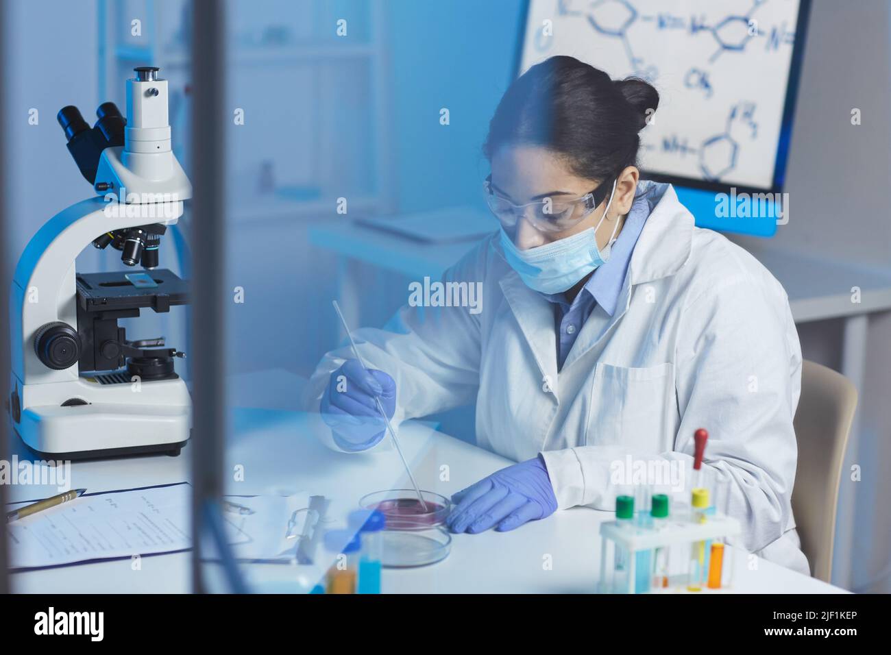 Serious young Indian female biologist in lab coat and surgical mask ...