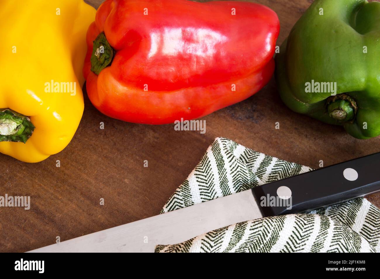 Three peppers of different colors on the table next to the knife and green striped fabric Stock Photo