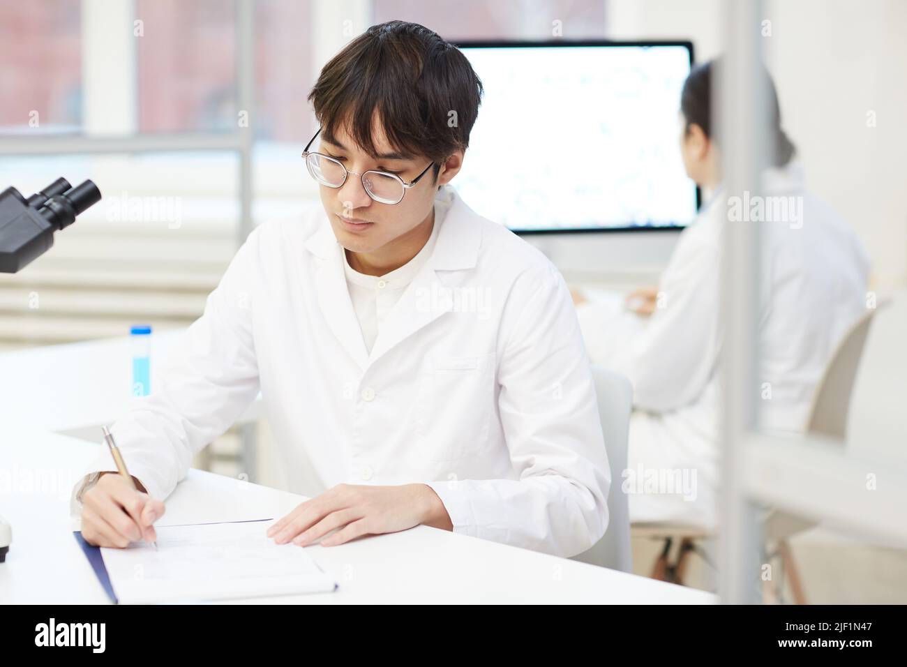 Serious concentrated young Asian lab technician in white coat and eyeglasses sitting at desk and making notes about laboratory research Stock Photo