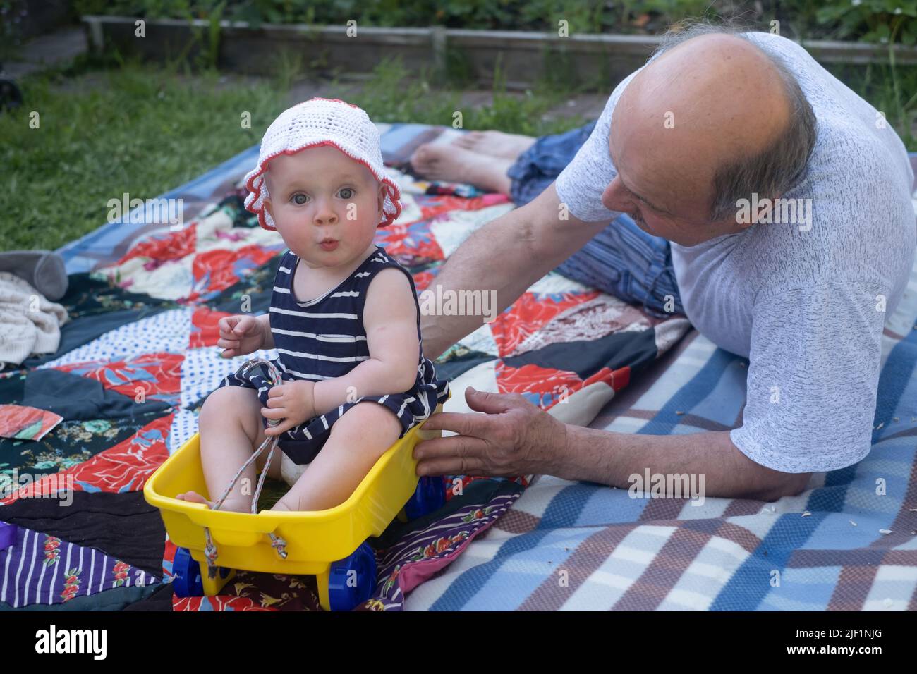 Senior grandfather playing with his grandchild on blanket outdoors pushing small cart. Stock Photo