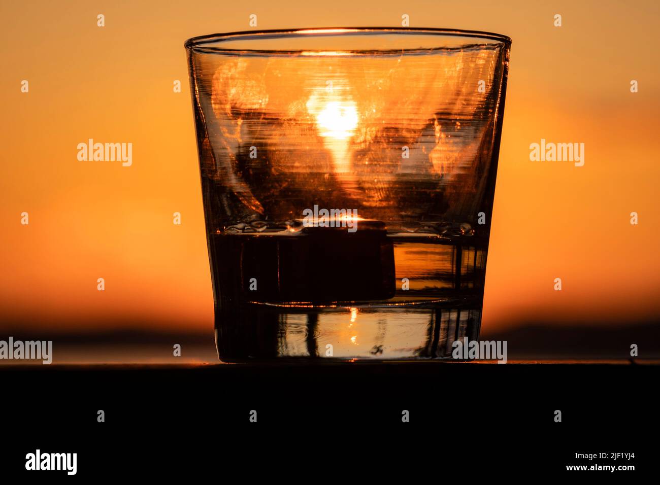 A glass of whisky sits on a railing while set against the setting sun