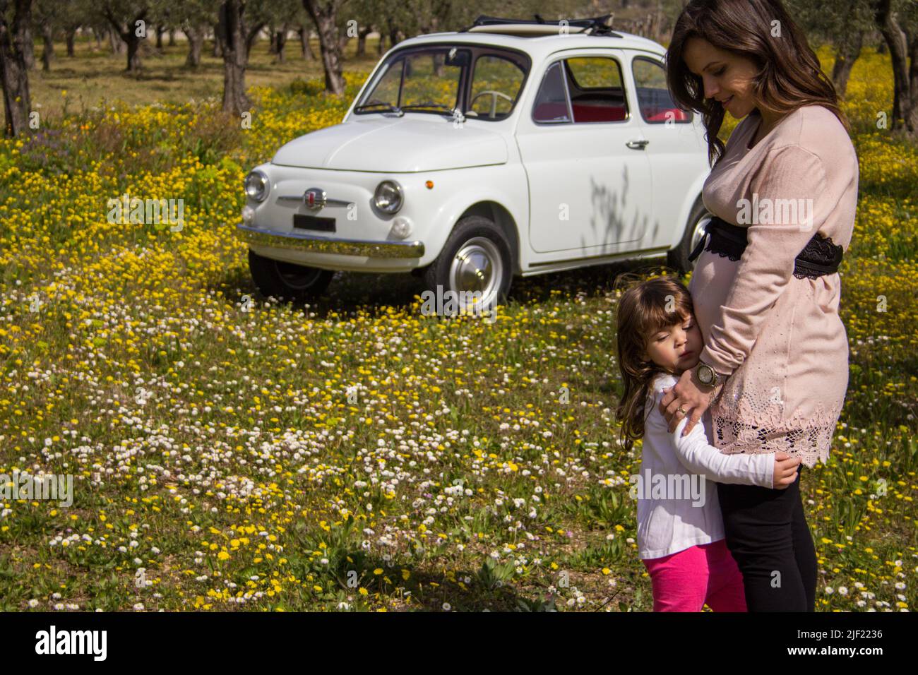 Image of a young expectant mother hugging her daughter in a flowery field with an old vintage car in the background. Vacation trip to Tuscany Italy Stock Photo