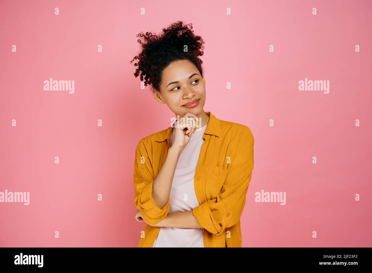 Positive african american curly young brunette woman, in orange casual shirt, looking away thoughtfully, coming up with an idea, contemplating a plan, daydreaming, stand on isolated pink background Stock Photo